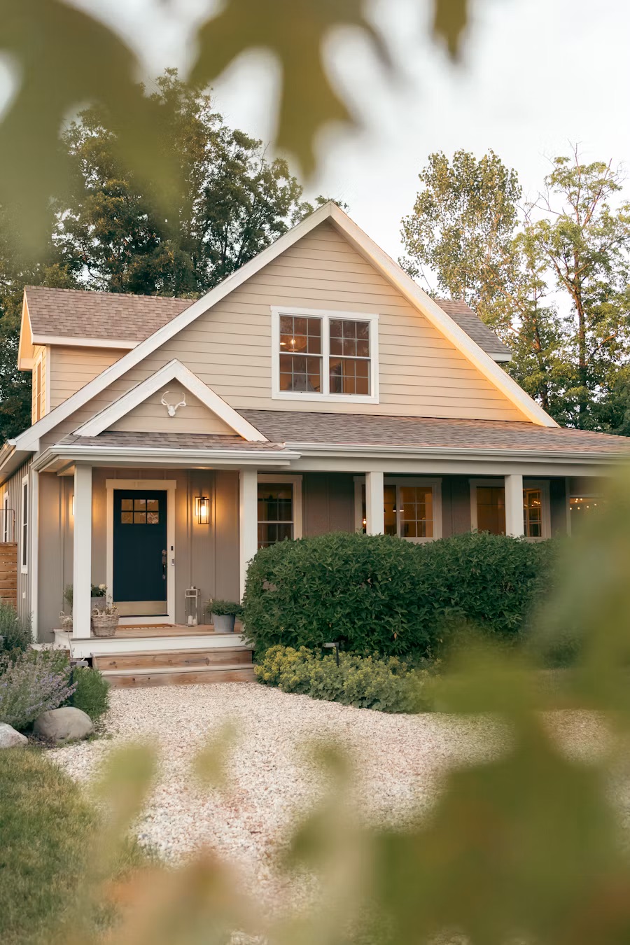 Front of a beige house with a covered porch, navy front door, lit lantern, and hedges, framed by leaves